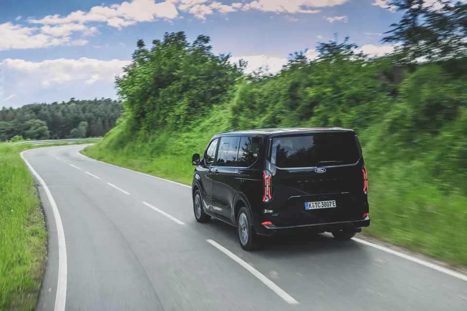 Rear view of a black Ford Tourneo travelling along a tree-lined road
